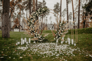 Arche de mariage circulaire moderne décorée de roses blanches, pivoines et herbes de pampas sur une pelouse parsemée de pétales.